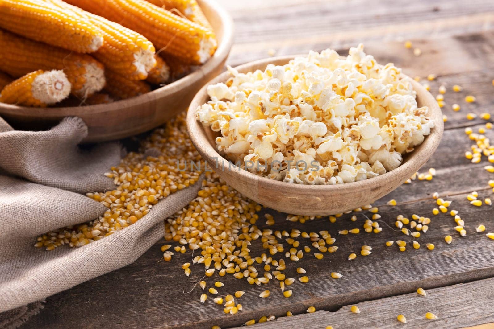 Wodden bowl of popcorn surrounded by popcorn kernels and popcorn on the cob