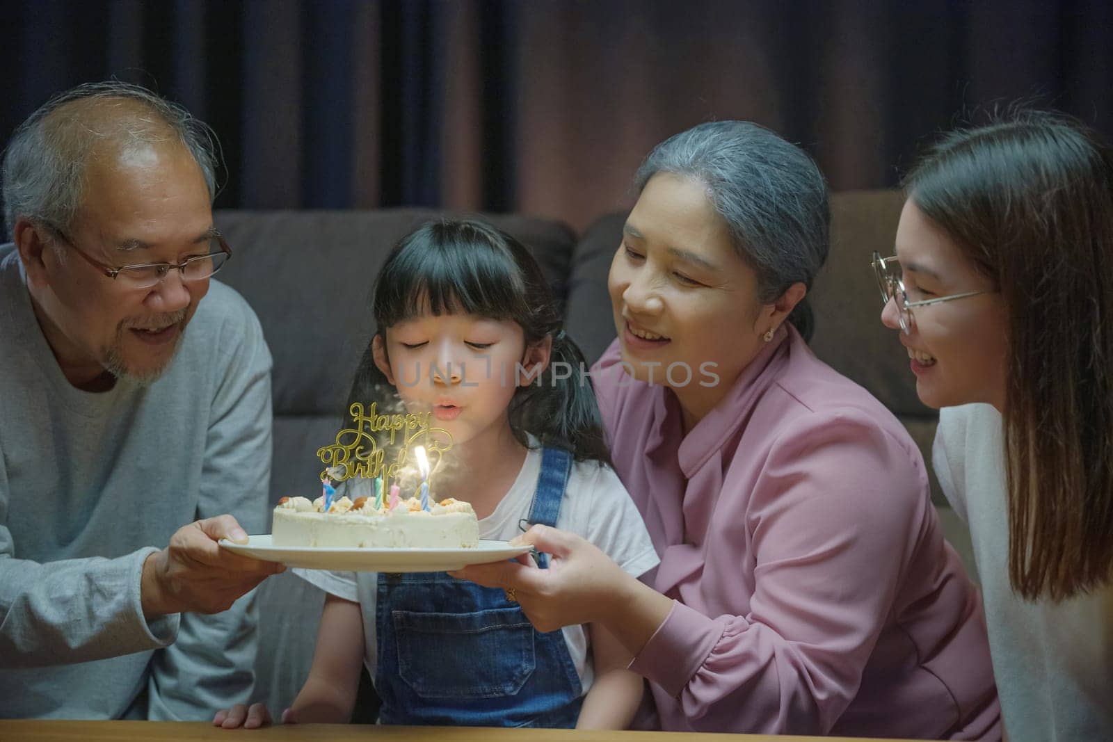 Happy Asian family grandparents and mother celebrating enjoy birthday party with cake of granddaughter Blowing candle at home in night time, Birthday surprise concept