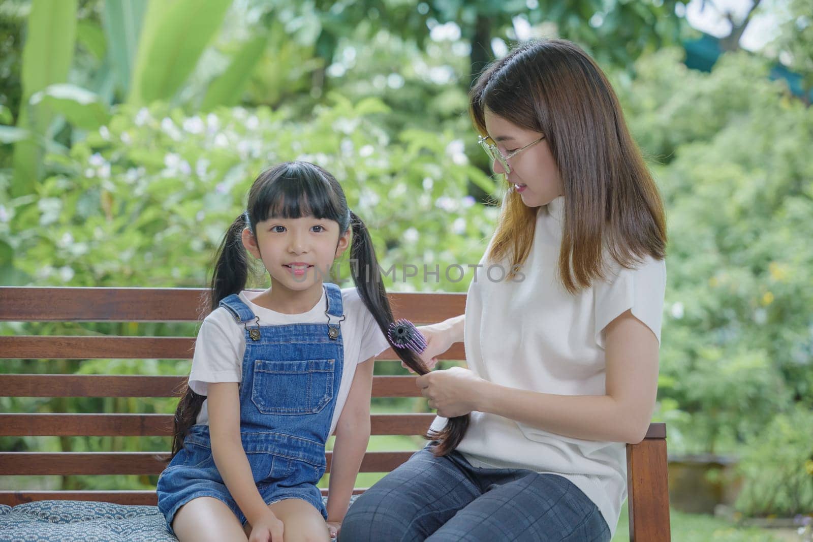 Happy family beautiful mother woman brushing her daughter hair while sitting outdoor at home, Asian smiling child schoolgirl with long dark hair and her mom in the morning, hairstyle care concept
