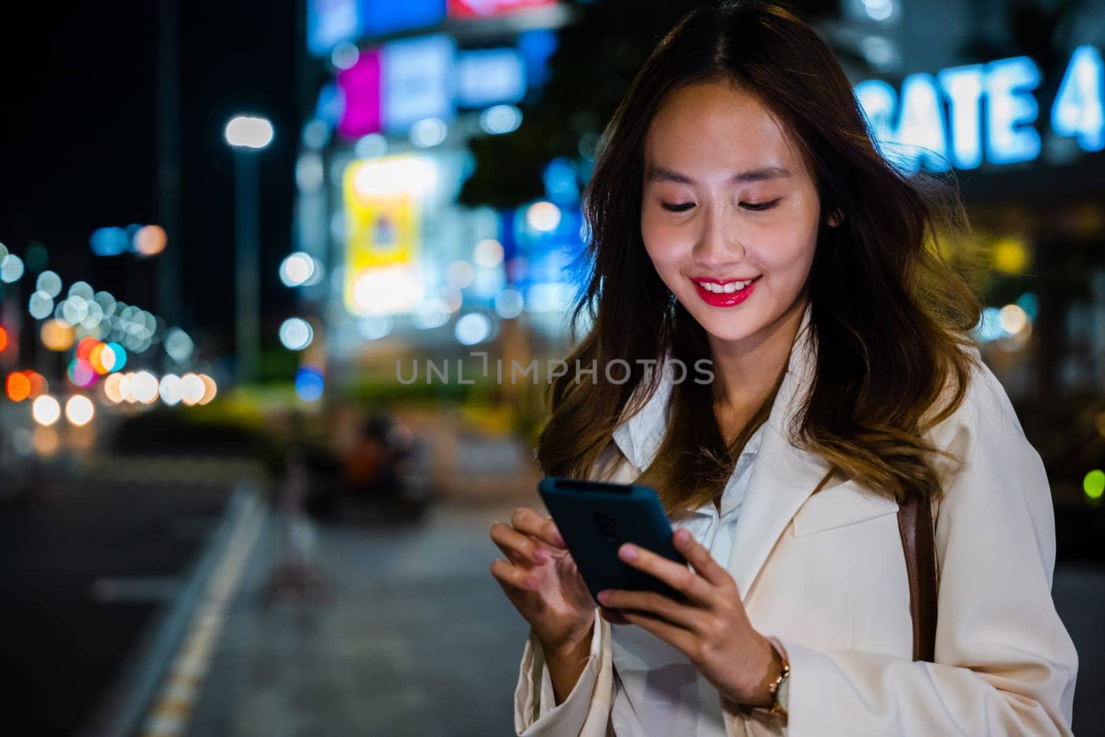 Business woman using mobile phone walking through night city street while waiting car to pick up home, Beautiful young smiling female texting work message on smartphone outside office, social media