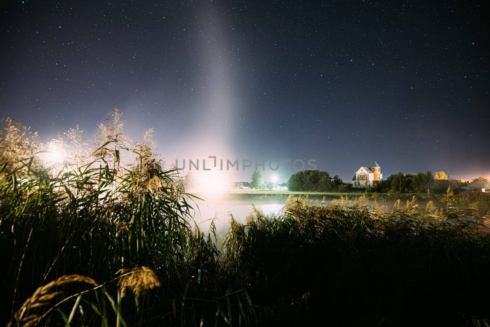 Evaporation Over River Lake Near Houses In Village. Night Starry Sky Above Misty Lake River Landscape At Night. Glowing Stars Above Summer Nature.
