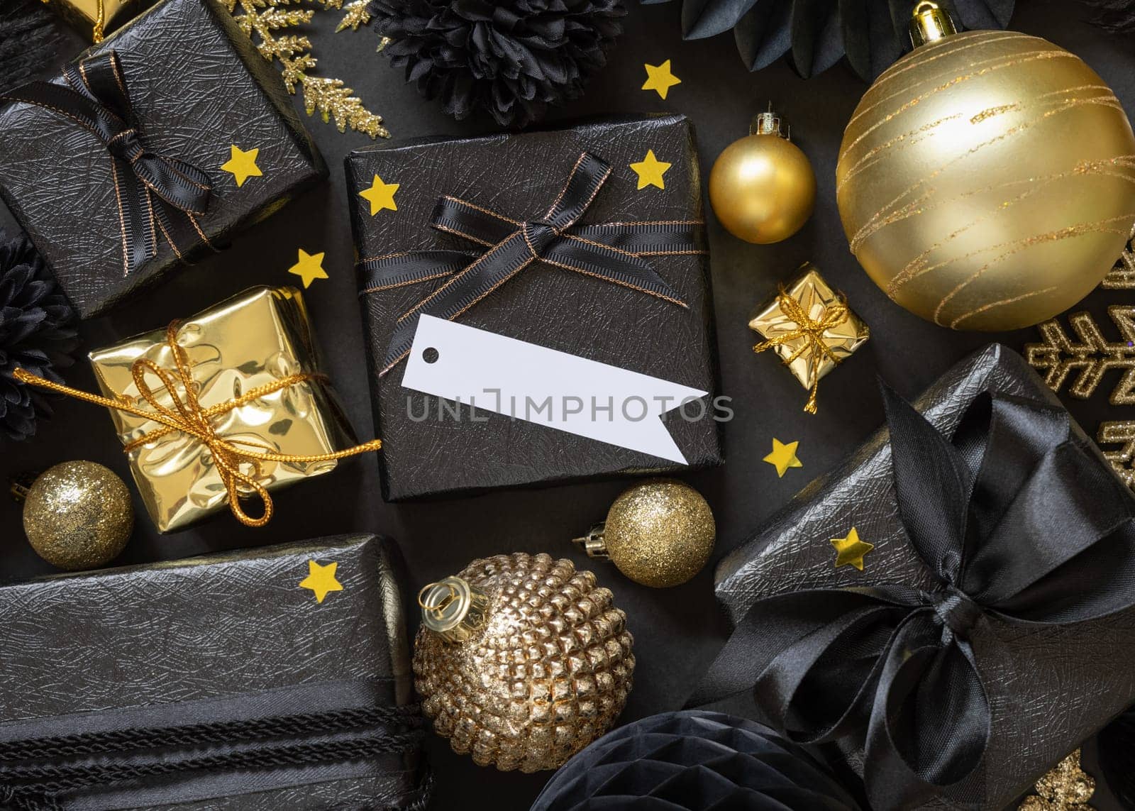 Black and golden Christmas Gift Boxes with a bow and ornaments close up, horizontal paper gift tag mockup, copy space. Dark winter composition with blank label card for Christmas, New Year, Birthday, Anniversary