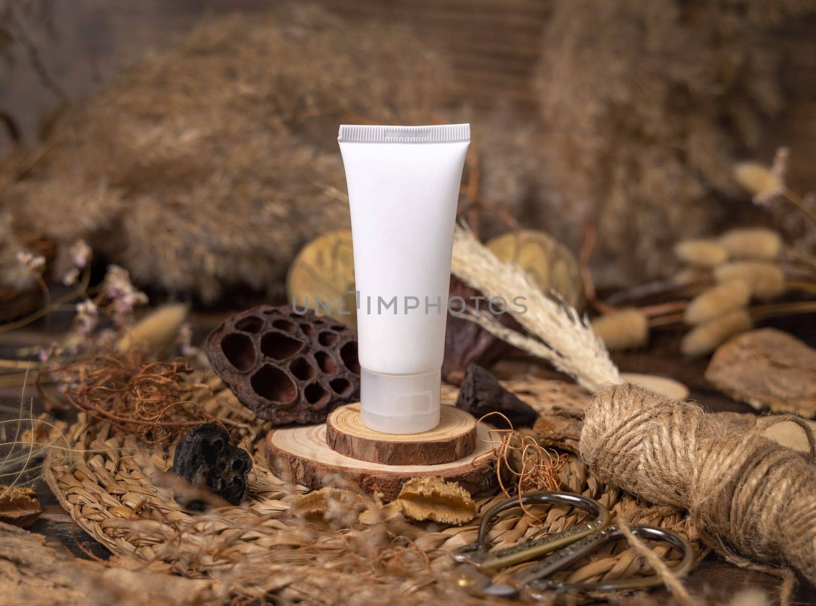 White plastic cosmetic tube on wood near natural decorations closeup, packaging mockup. Eco friendly skincare product, lotion or cream. Bohemian composition with dried leaves and flowers.