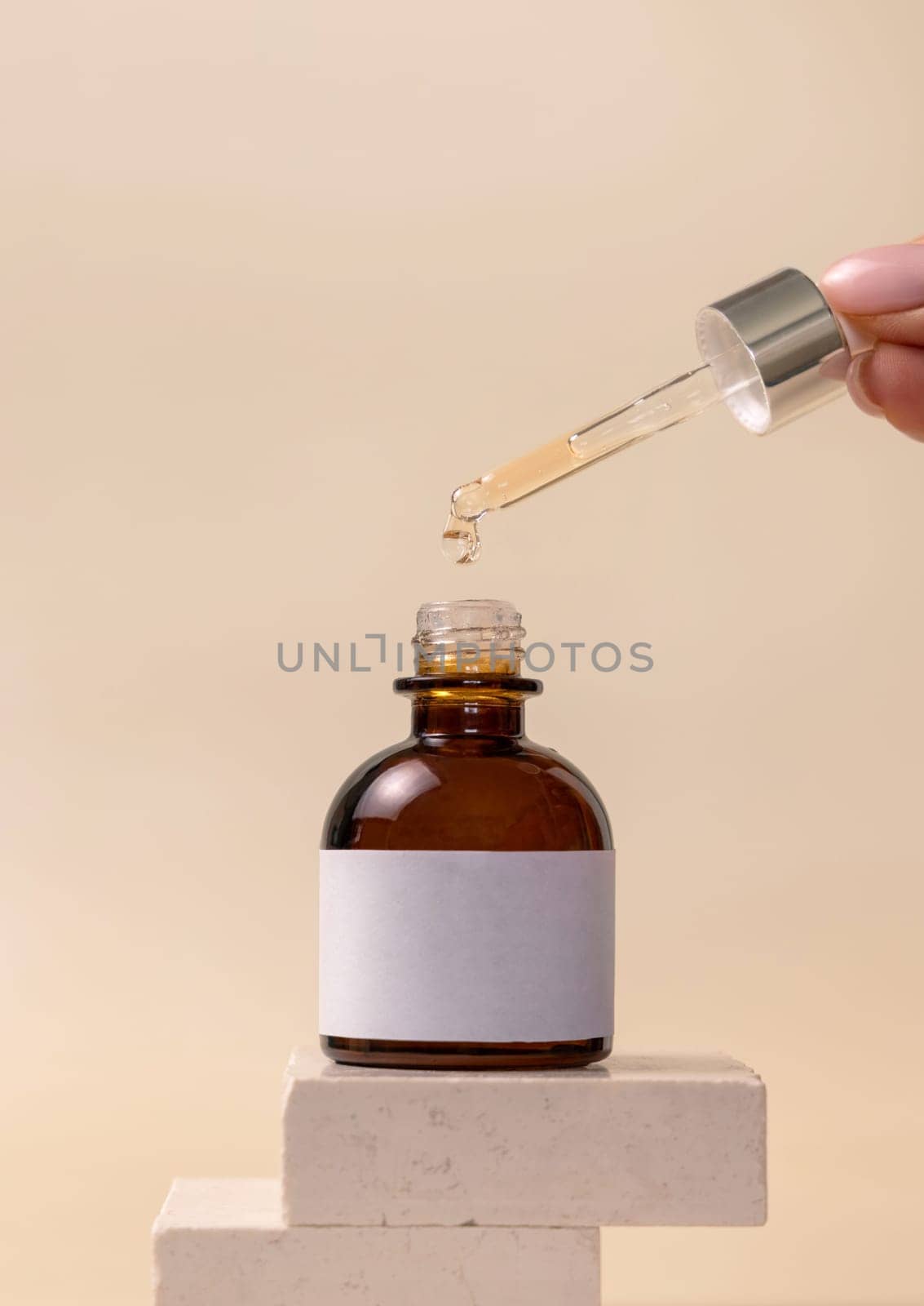 Hand holding pipette with drop falling down into opened dropper bottle on stones on light beige, close up, cosmetic label mockup. Natural skincare product, serum or essential oil.