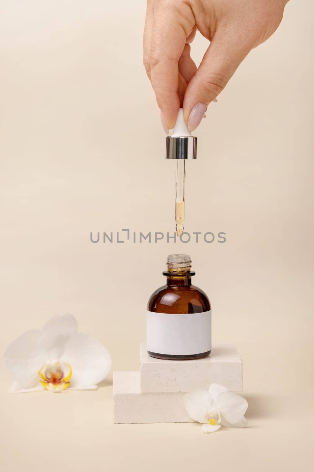 Hand holding pipette over opened cosmetic dropper bottle near white orchid flowers and stones on light beige, close up, mockup. Natural skincare product, serum or essential oil.