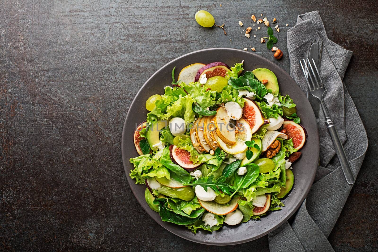 Fresh green leafy salad with mozzarella cheese, fruits and nuts on dark background close up. Concept for a tasty and healthy meal