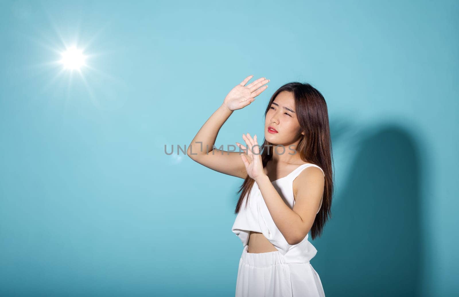 Asian young woman hand up cover face protect sun light and UV smile in studio shot isolated on blue background, female raising arm and hand in protection and block sunshine, sunscreen cream advertise