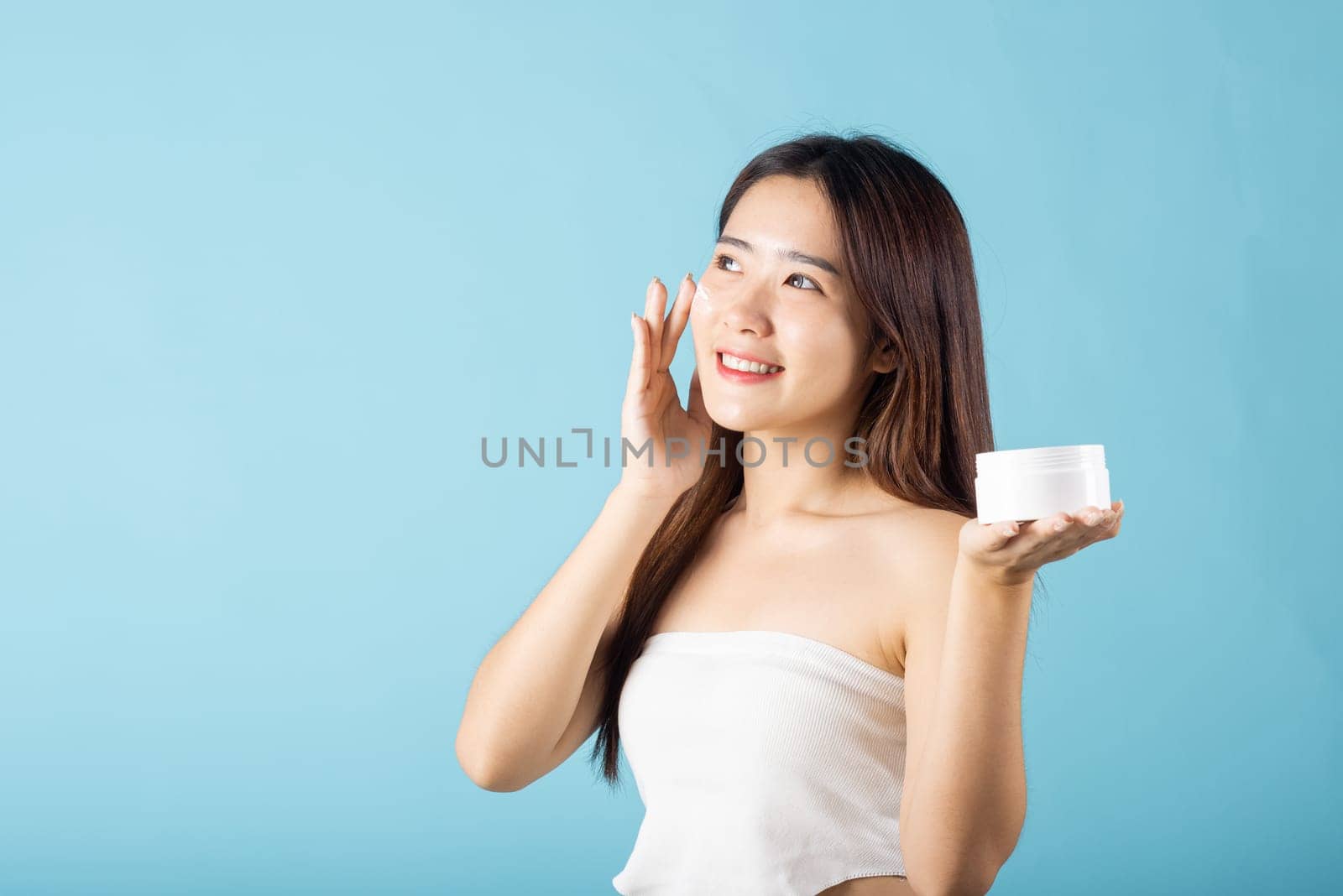 Asian young woman are not very beautiful hold jar of skin cream for face and apply anti-aging moisturizer studio isolated on blue background, Happy female pointing finger her face, pursuit of beauty