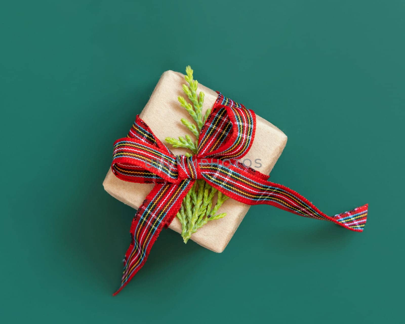 Gift box wrapped in karft paper with red ribbon bow and juniper leaf on green top view. Christmas or New Year Holiday atmosphere flat lay