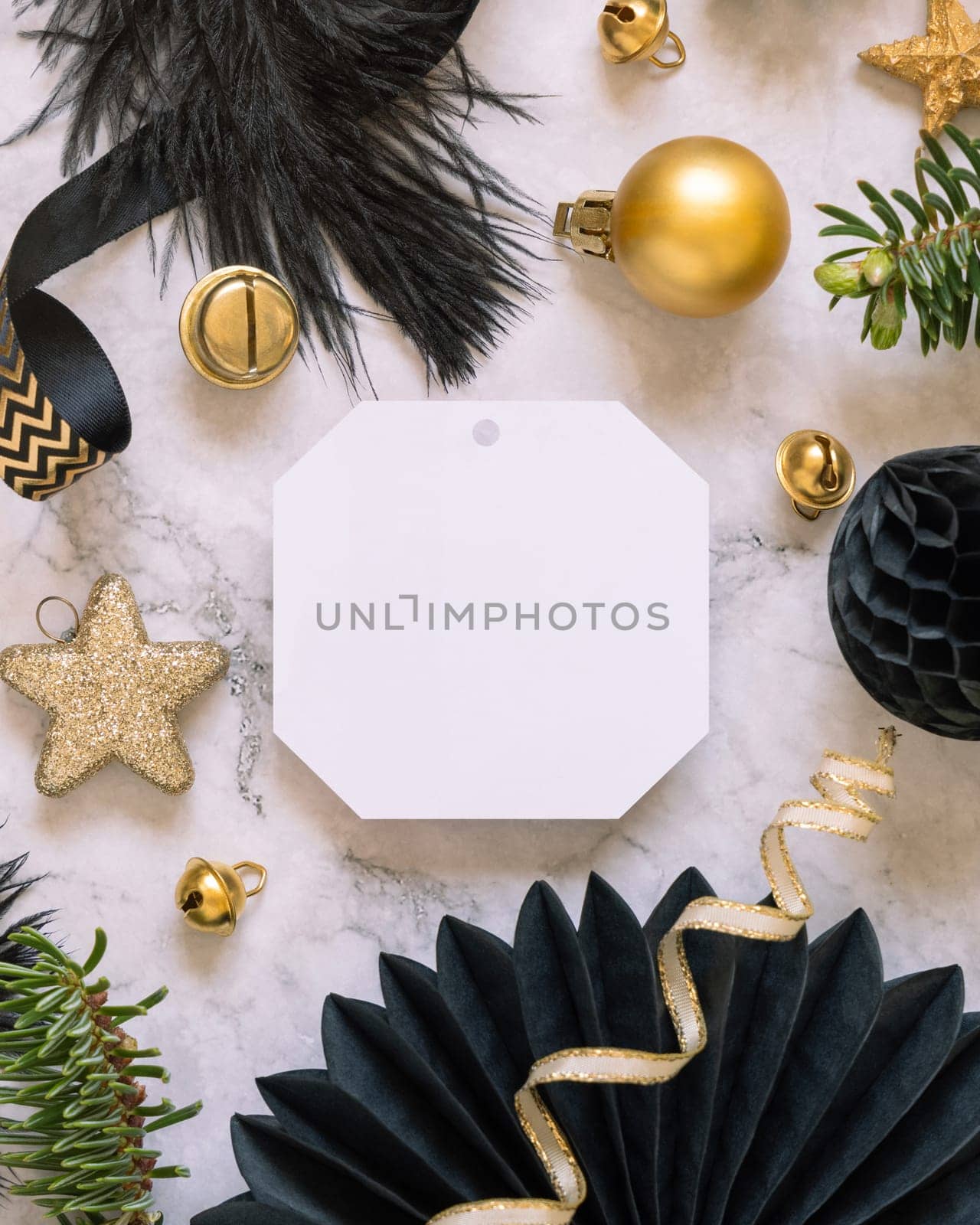 Paper gift tag between fir tree branches and black and golden Christmas decorations on a white marble table top view. Mockup, copy space. Winter holiday flat lay with blank label