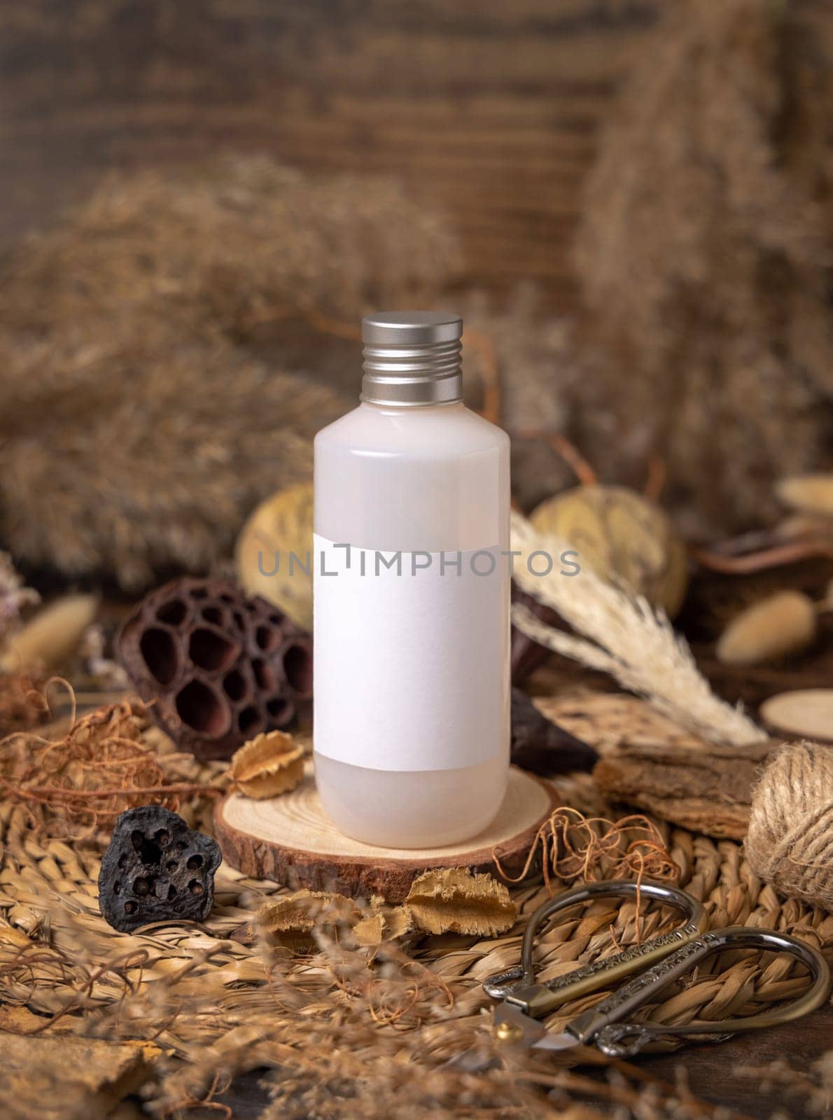 Cosmetic bottle with blank label on wood near natural decorations close up, package mockup. Eco friendly skincare product, lotion or cream. Bohemian composition with dried leaves and flowers.