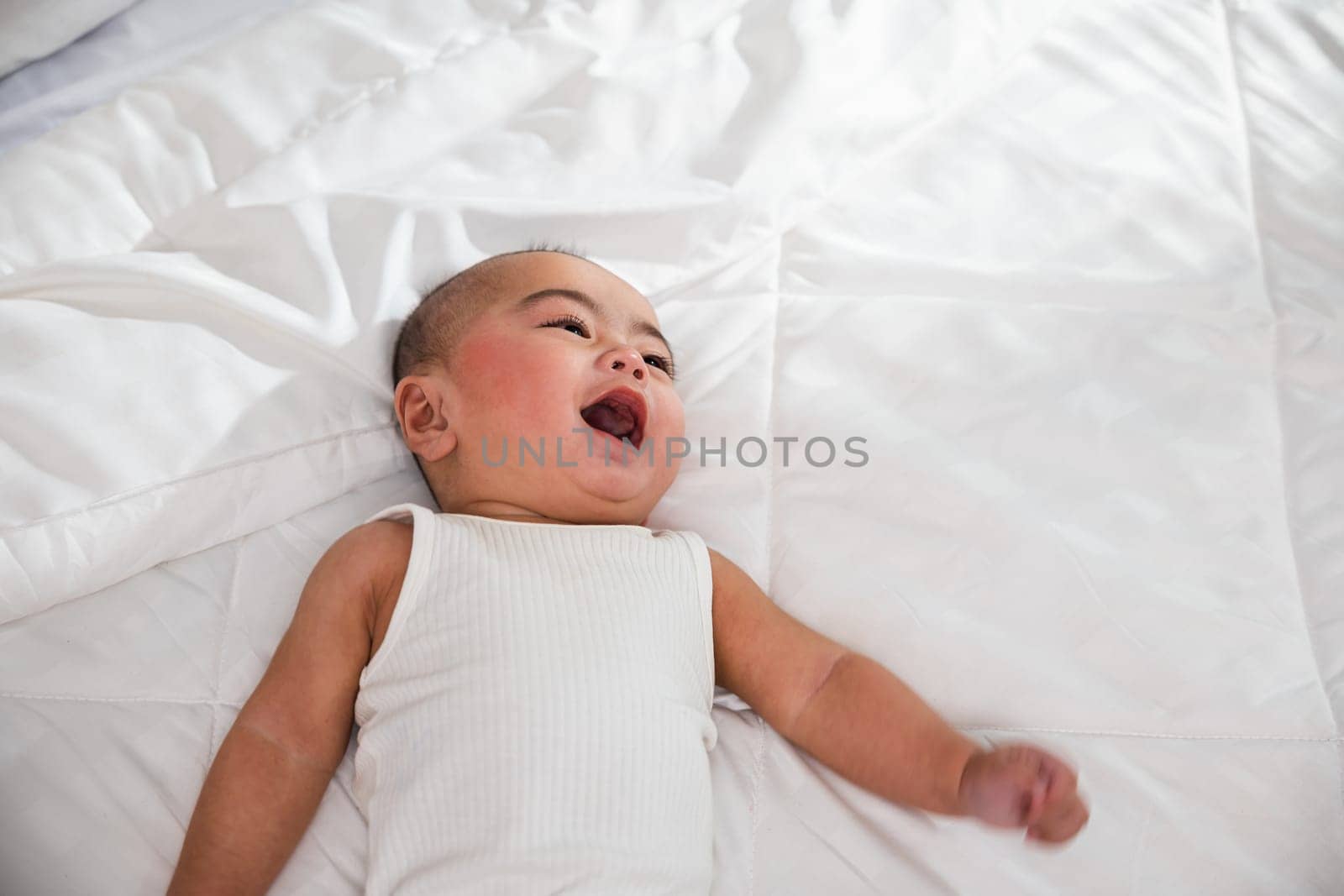 Asian cute little baby on bed under soft blanket, baby wearing bodysuit lying on white beedsheets in bedroom, smiling infant child at home