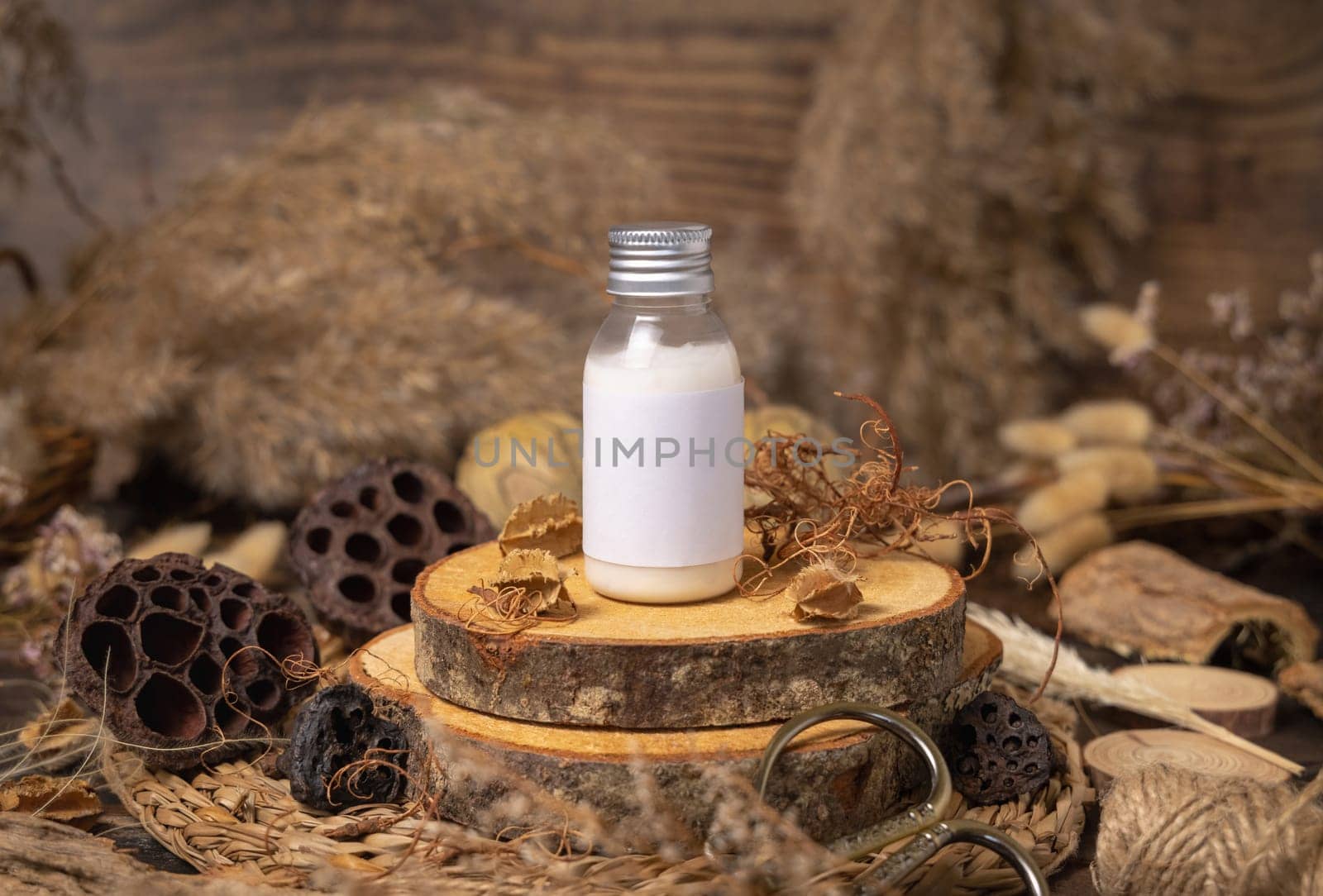 Cosmetic bottle with blank label on wood near natural decorations close up, package mockup. Eco friendly skincare product, lotion or cream. Bohemian composition with dried leaves and flowers.