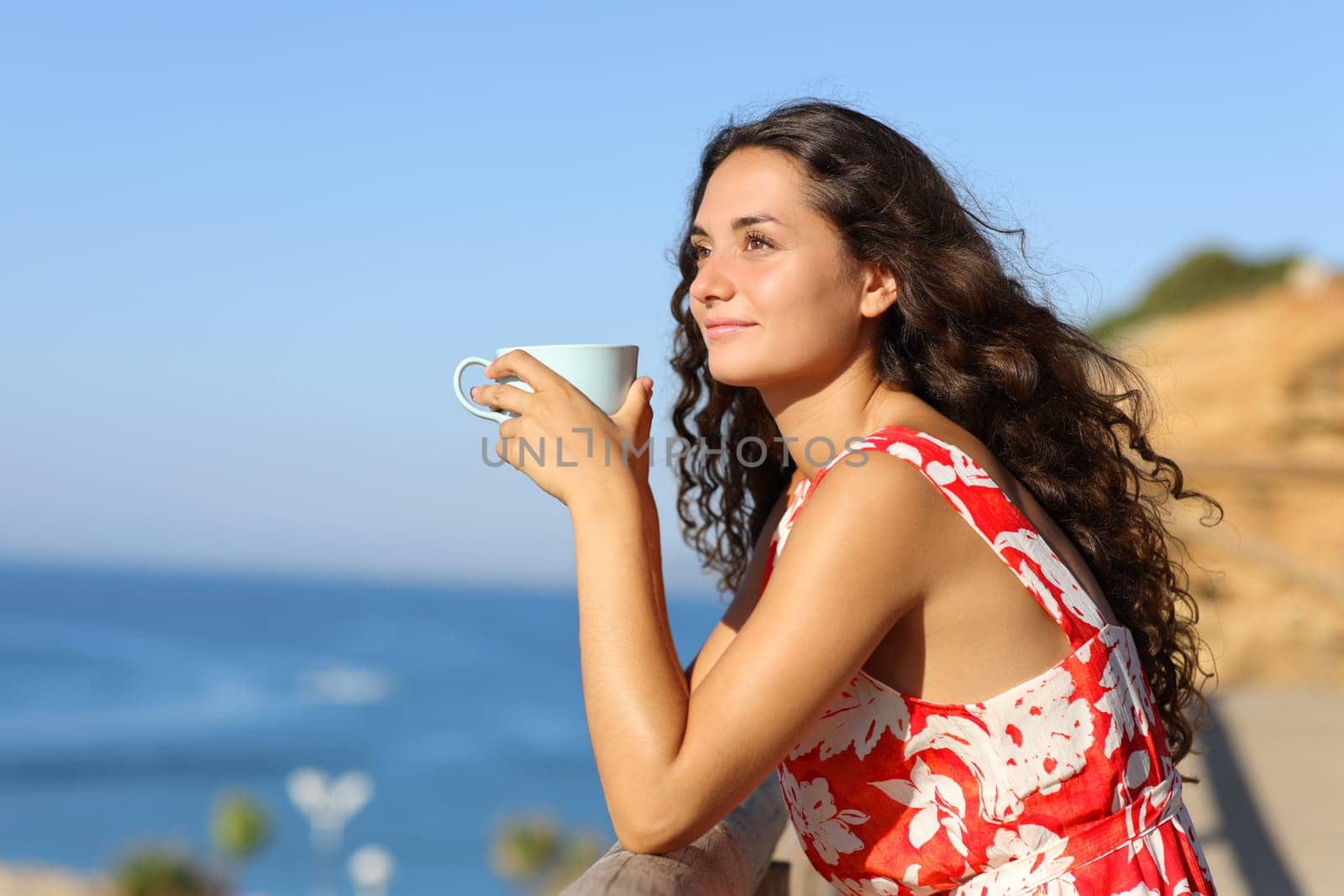 Woman in red drinking coffee on the beach looking away a sunny day