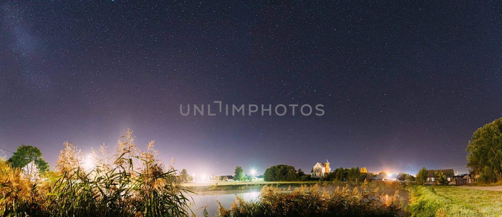 Panorama Of Evaporation Over River Lake Near Houses In Village. Night Starry Sky Above Lake River With Bright Stars And Meteoric Track Trail. Glowing Stars Above Summer Nature.