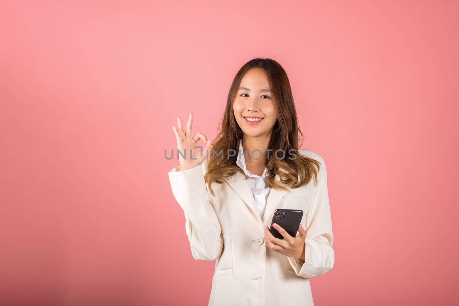 Happy Asian portrait beautiful cute young woman excited holding mobile phone and gesturing ok sign for agree, studio shot isolated on pink background, Thai female making finger symbol on smartphone