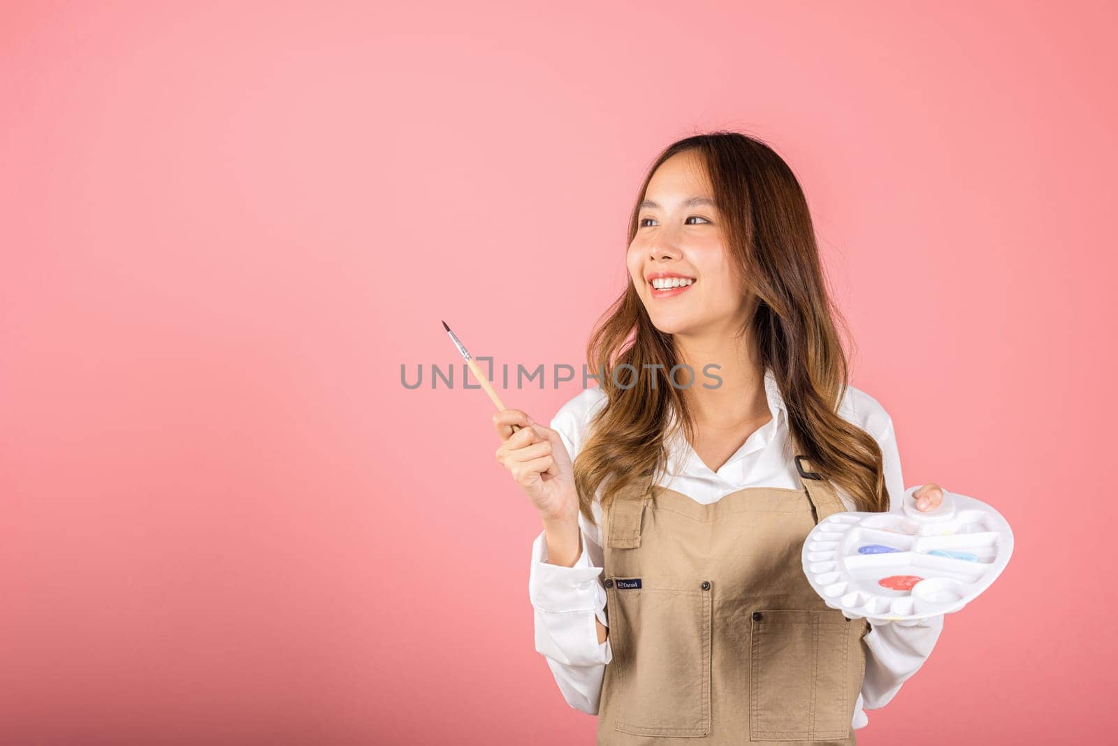 Happy female painting using paintbrush and palette with colors, Asian beautiful young woman artist holding brush and paint palette, studio shot isolated on pink background, Paintings and art equipment