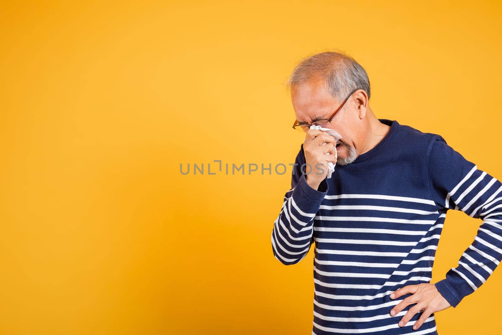 Asian elder man cold having flu and sneezing from sickness virus problem use tissue studio shot isolated on yellow background, Portrait senior old man blowing nose with tissue, pensioner unwell