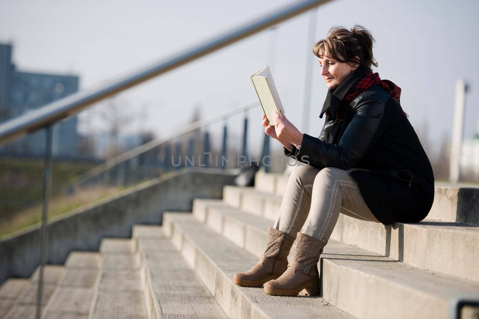 Side full body view of a mature woman in black leather jacket on a concrete staircase in a book reading against a fuzzy urban background