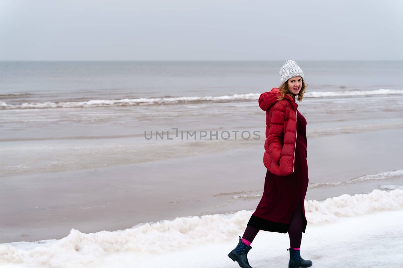 A woman in a red jacket and a red knitted dress walks in winter along the sea.