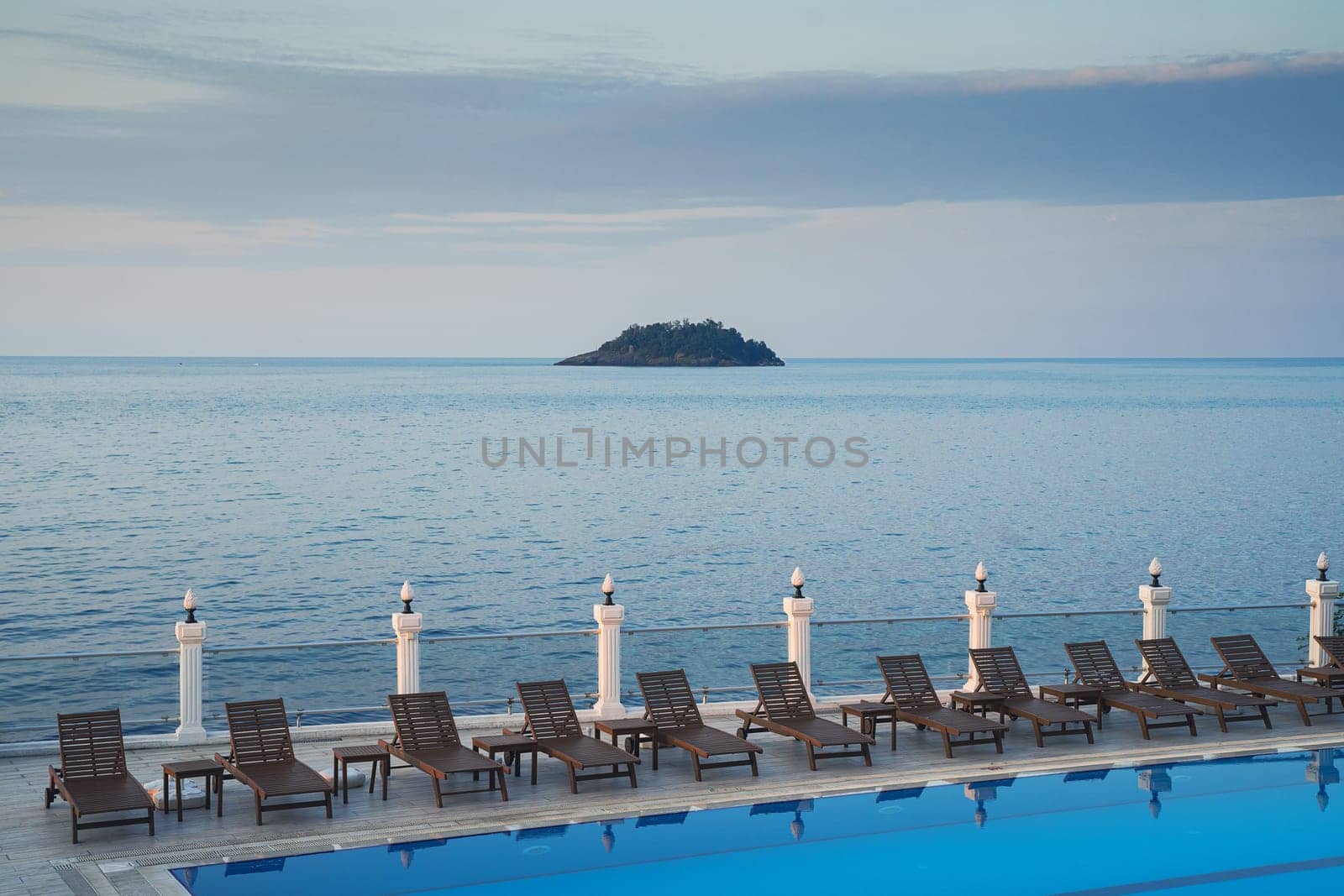 wooden sunlounger chairs near a swimming pool with blue water with sea and a small island in the background at sunset