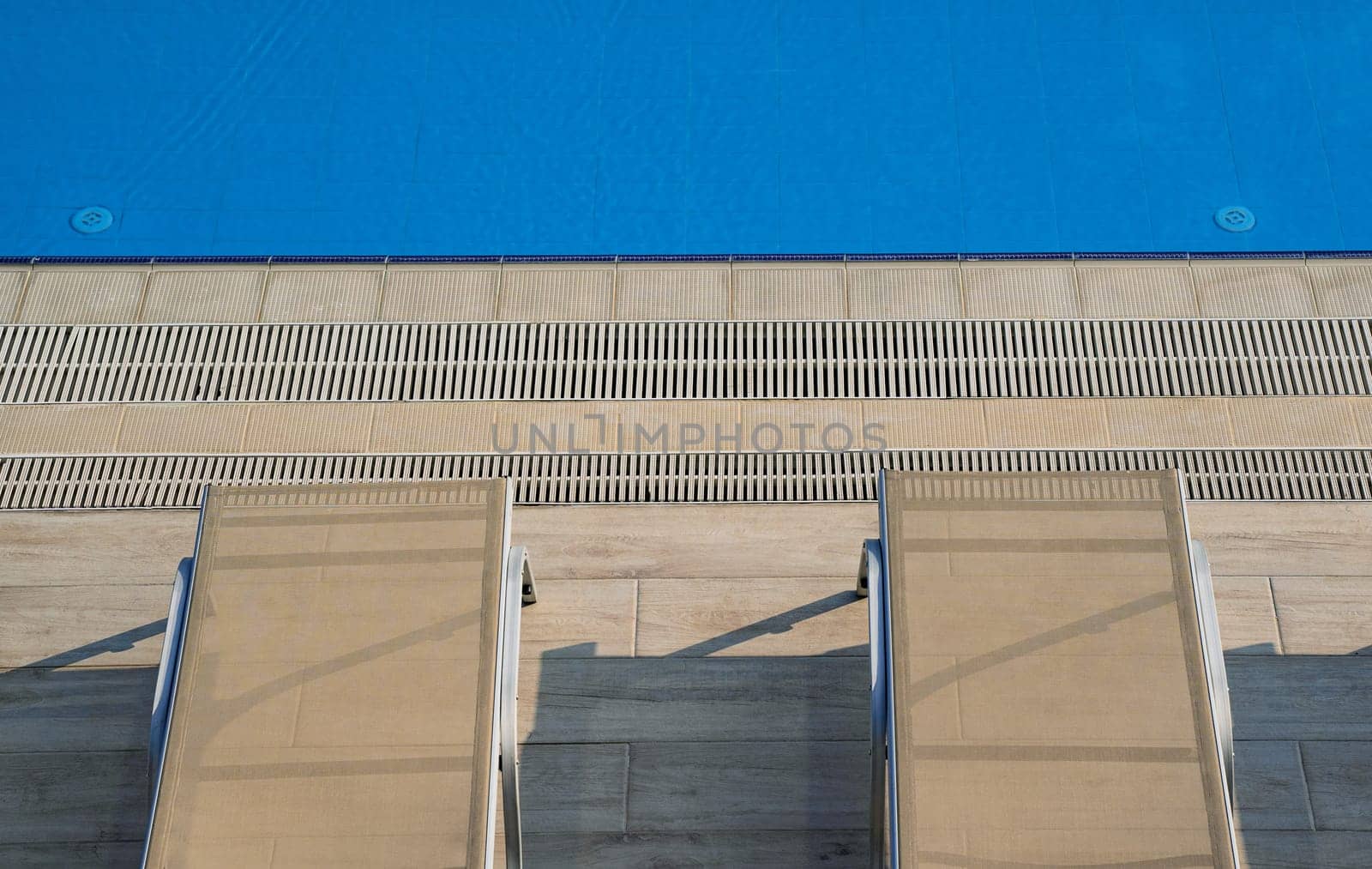 top view closeup of row of sunlounger chairs near a outdoor swimming pool with blue water and marble deck patio in the morning sun
