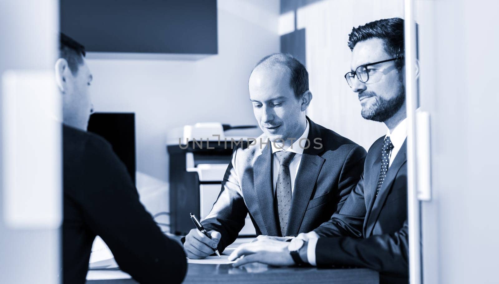 Team of confident successful business people reviewing and signing a contract to seal the deal at business meeting in modern corporate office. Business concept. Greyscale blue toned image.