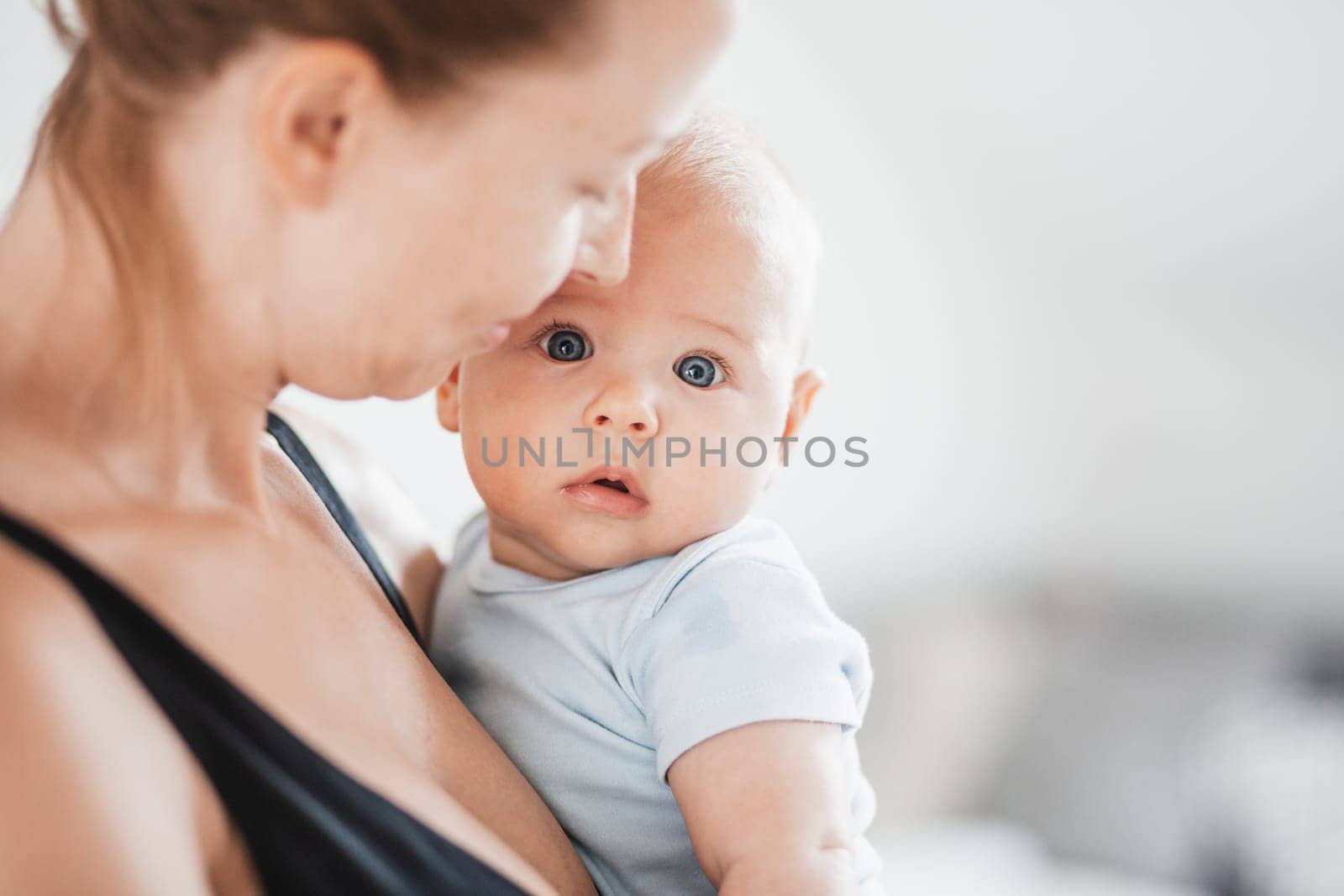 Portrait of sweet baby resting in mothers arms, looking at camera. New mom holding and cuddling little kid, embracing child with tenderness, love, care. Motherhood concept.