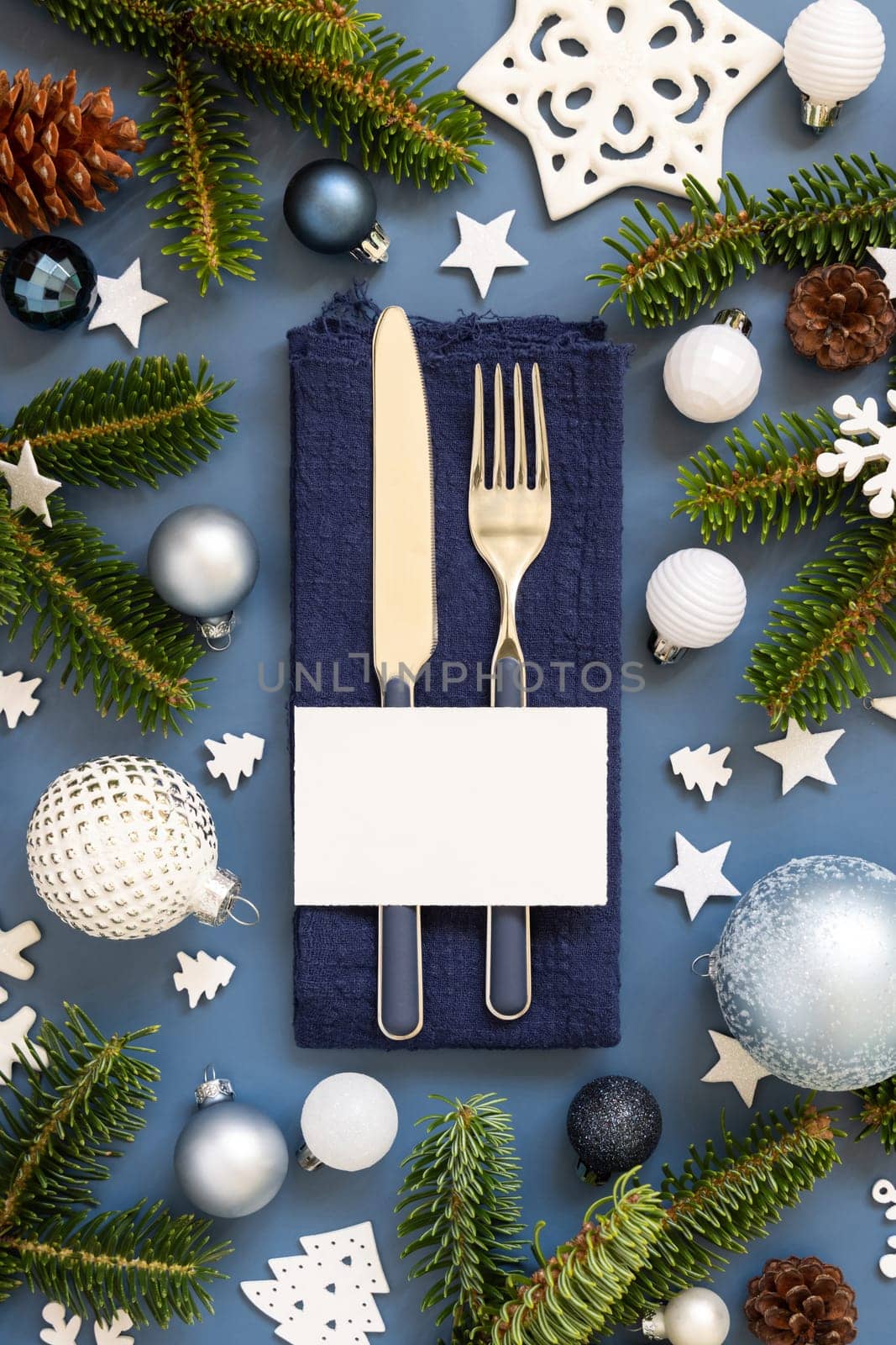 Blue and white Christmas table setting with blank card near decorations and fir tree branches top view. Winter table with fork, knife and plate. Winter New Year or Christmas dinner atmosphere