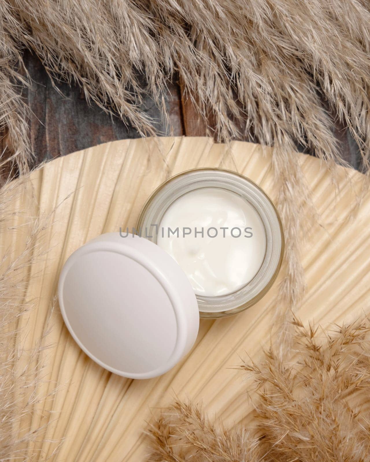 Opened cream jar with a blank lid on dried palm leaf near pampas grass top view, mockup. Skincare beauty product. Bohemian eco friendly cosmetics flat lay with dried leaves on wood.
