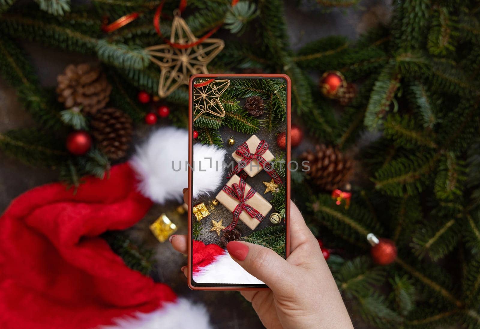 Woman's hand taking a mobile photo of christmas composition with Santa Claus hat, fir branches and holiday decorations top view. Blogger preparing content for social media using cell phone.