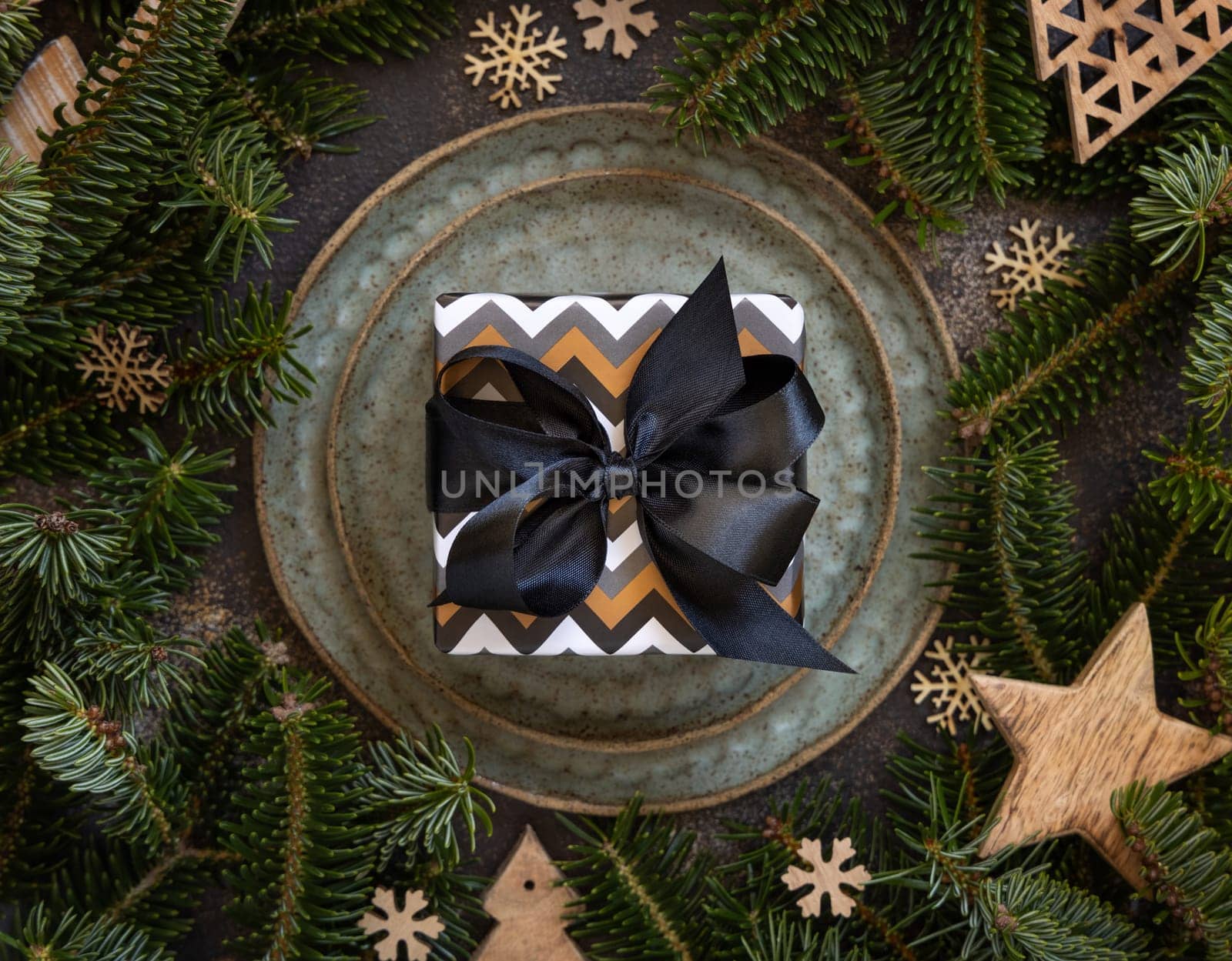 Wrapped present on Festive table near fir tree branches and Rustic Christmas decorations top view. Christmas or New Year table place with gift box on green plates on dark table flat lay
