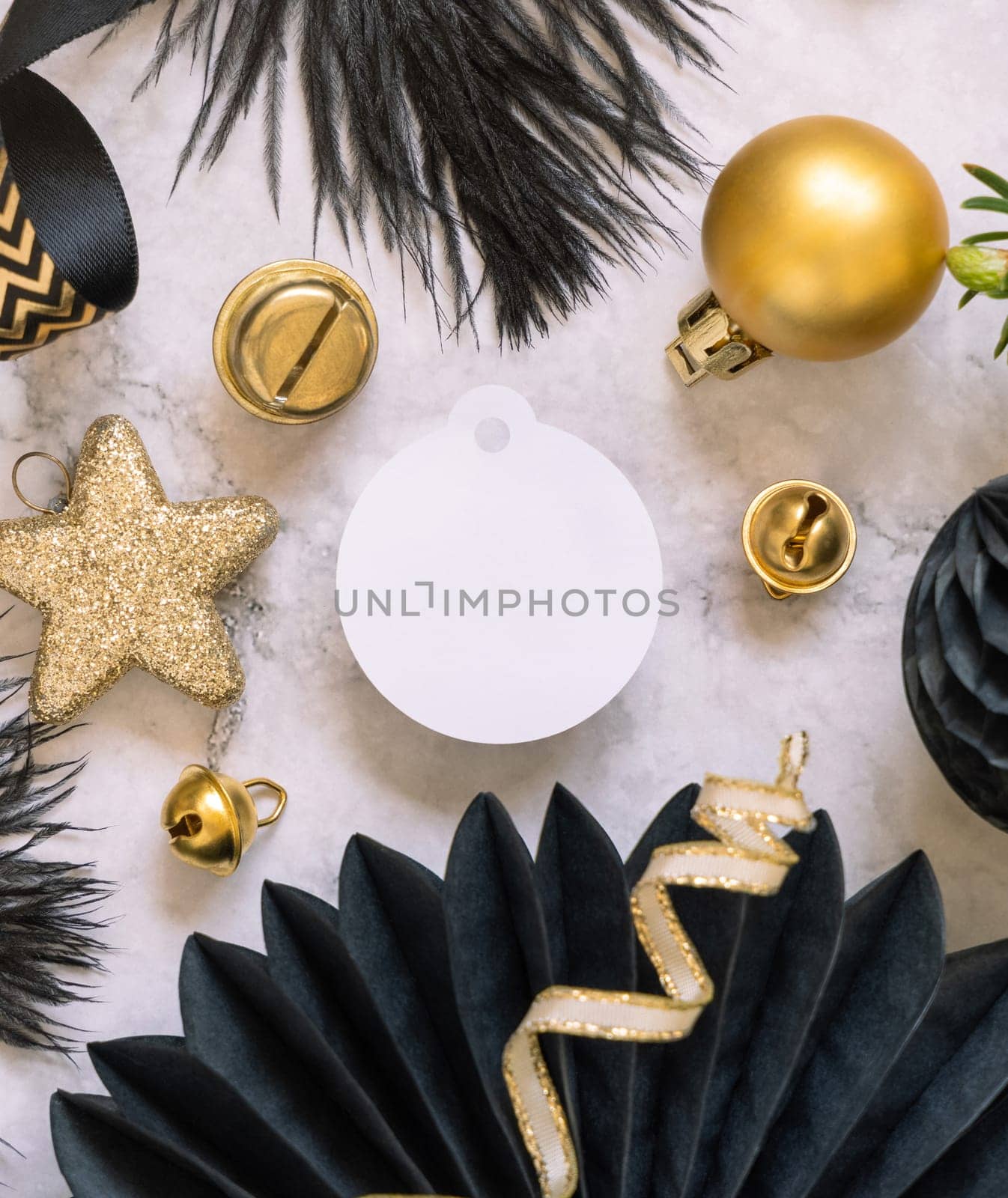 Paper round gift tag between fir tree branches and black and golden Christmas decorations on a white marble table top view. Mockup, copy space. Winter holiday flat lay with blank label