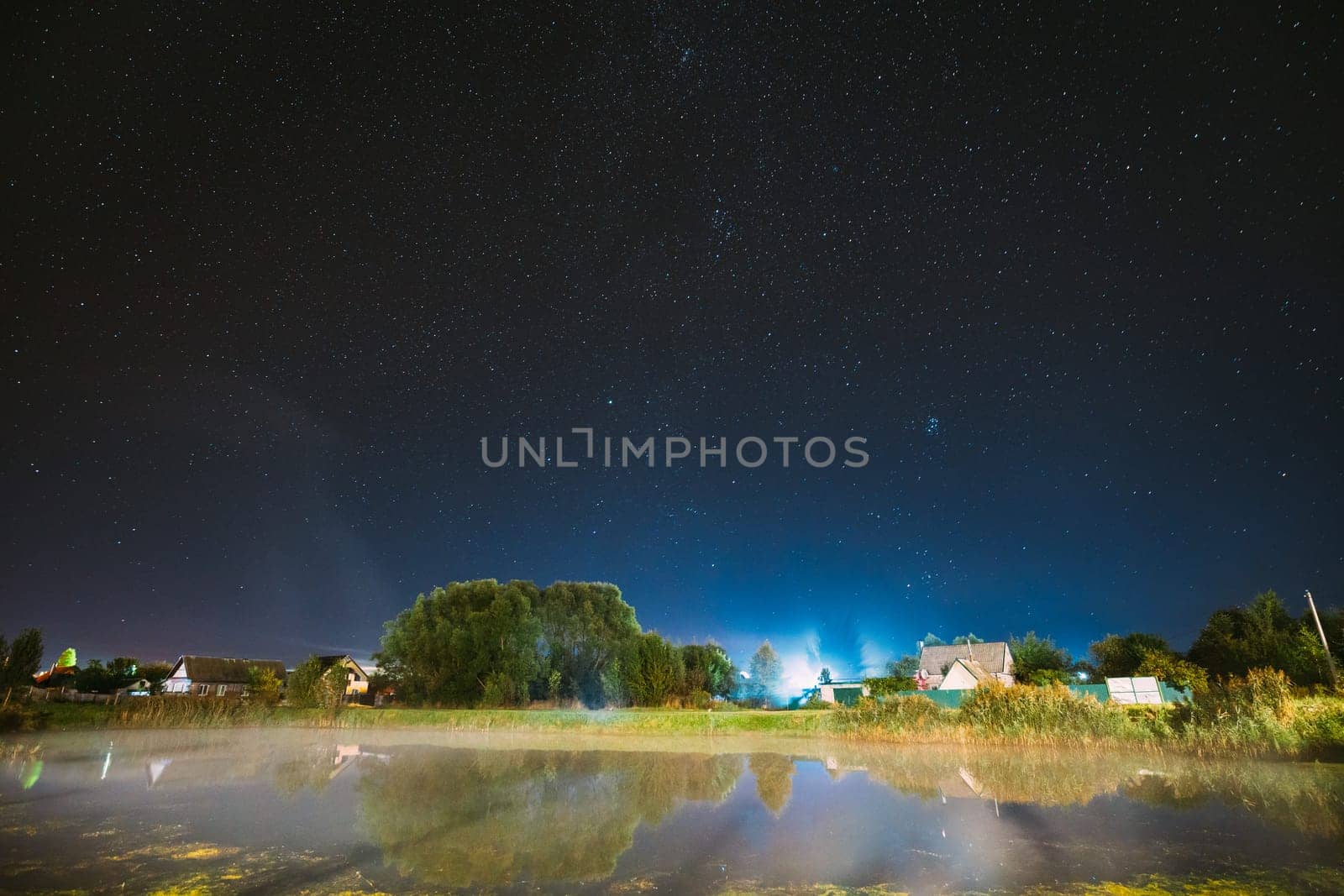 Evaporation Over River Lake Near Houses In Village. Milky Way Galaxy In Night Starry Sky Above Lake River Landscape At Night. Glowing Stars Above Summer Nature
