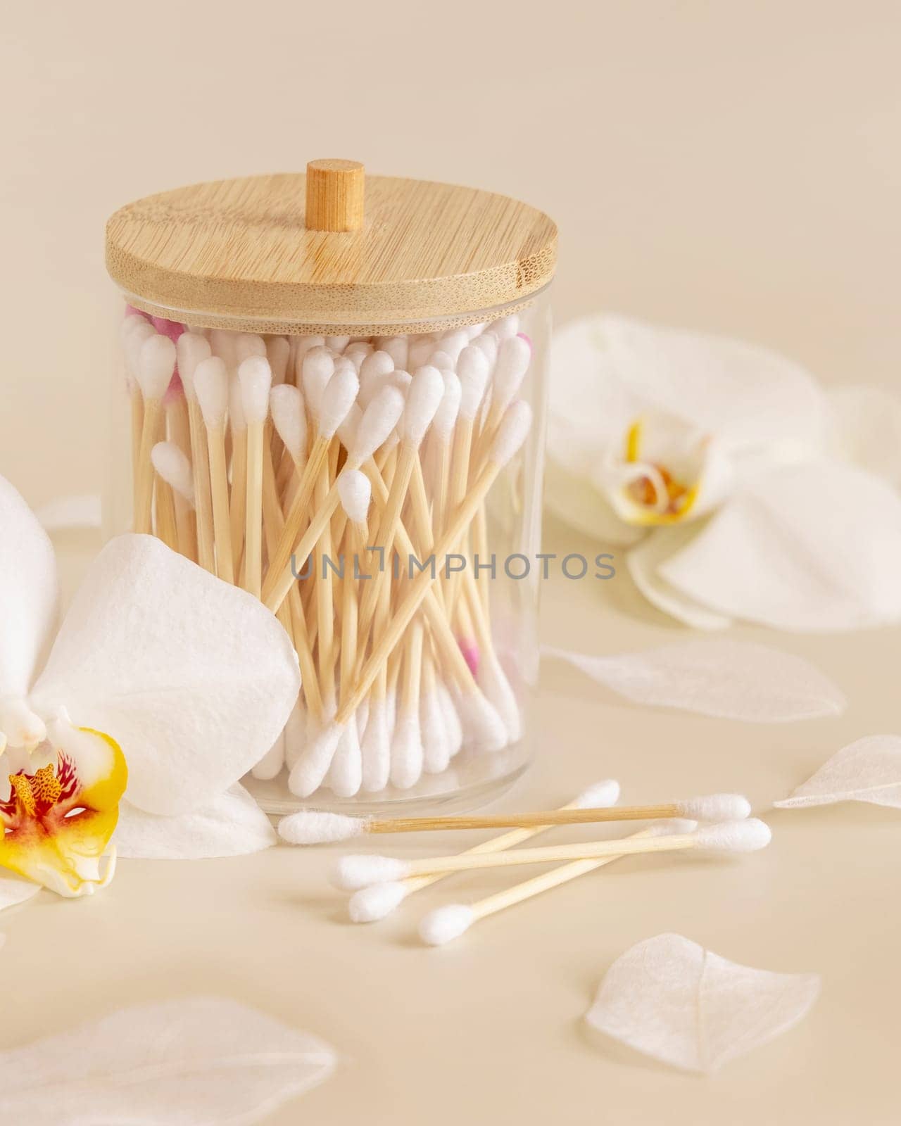 Bamboo cotton buds in a jar near white orchid flowers on light beige, close up. Organic skincare product for everyday beauty routine. Eco friendly Biodegradable cotton swabs..