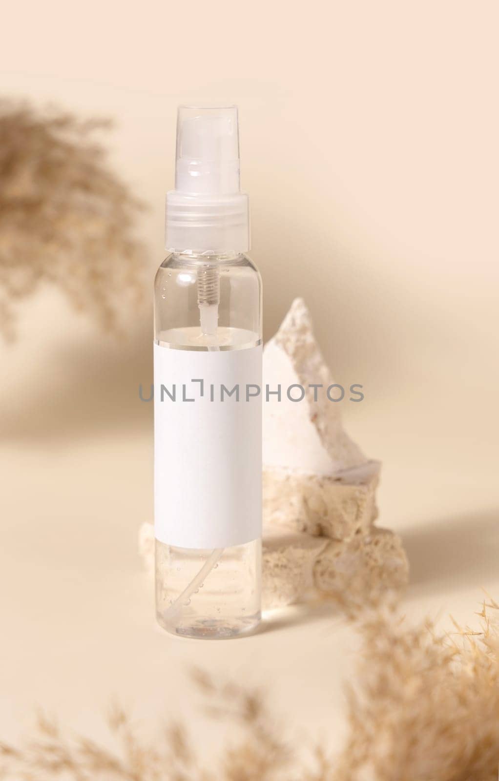 Transparent glass cosmetic bottle with blank label on stones near pampas grass on light yellow, close up, mockup. Skincare beauty product. Natural eco friendly cosmetics