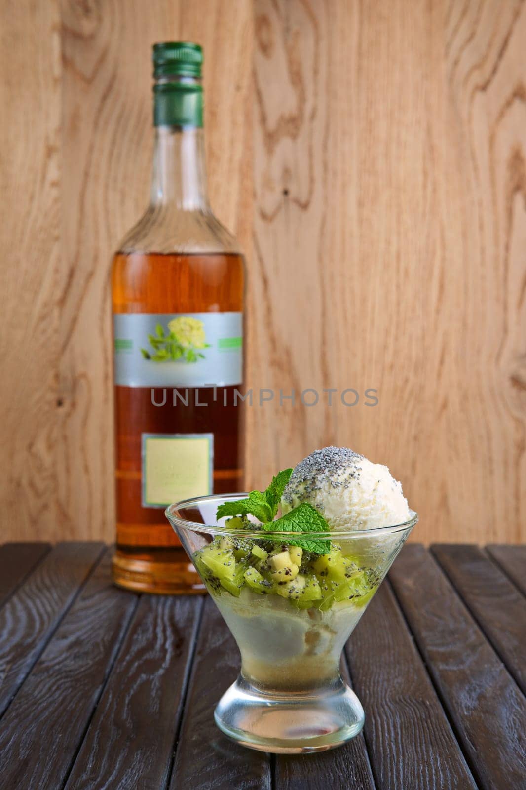A cup of apple ice cream decorated with kiwi, mint leaves and poppy. Bottle of syrup on background.