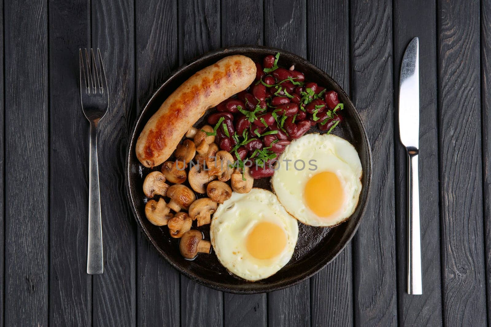 Cast-iron frying pan with fried eggs, sausage, mushroom and beans on wooden table. Top view.