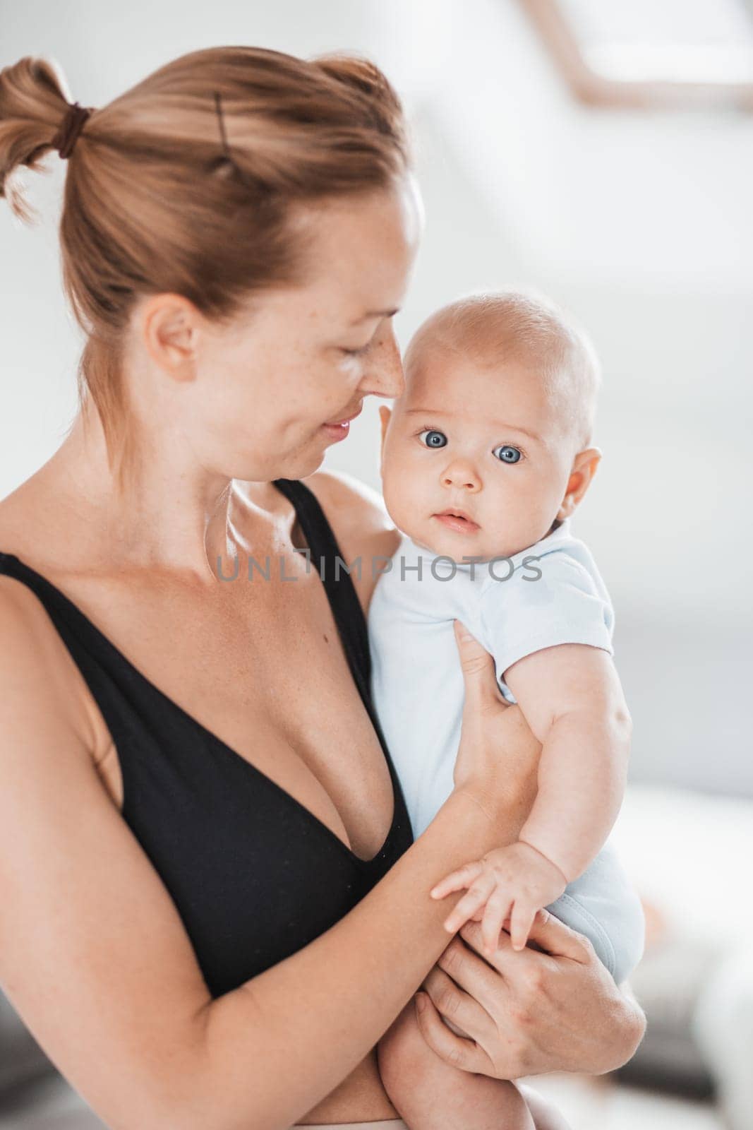 Portrait of sweet baby resting in mothers arms, looking at camera. New mom holding and cuddling little kid, embracing child with tenderness, love, care. Motherhood concept.