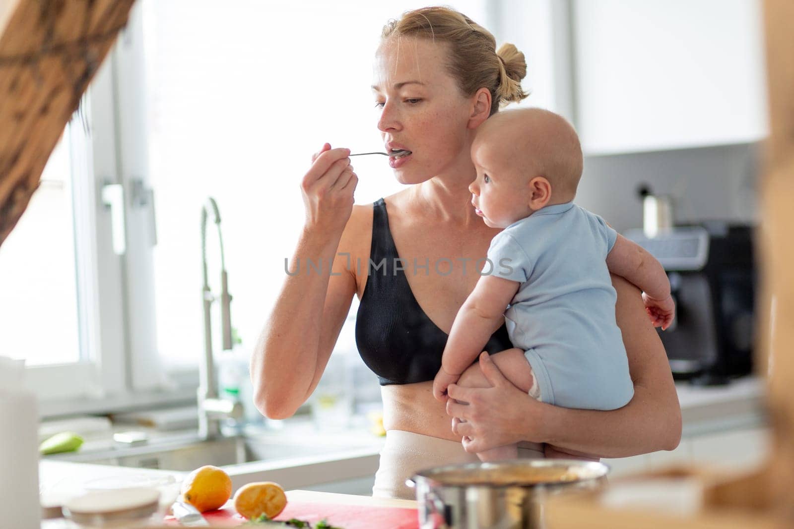 Woman cooking and tasing food while holding four months old baby boy in her hands.