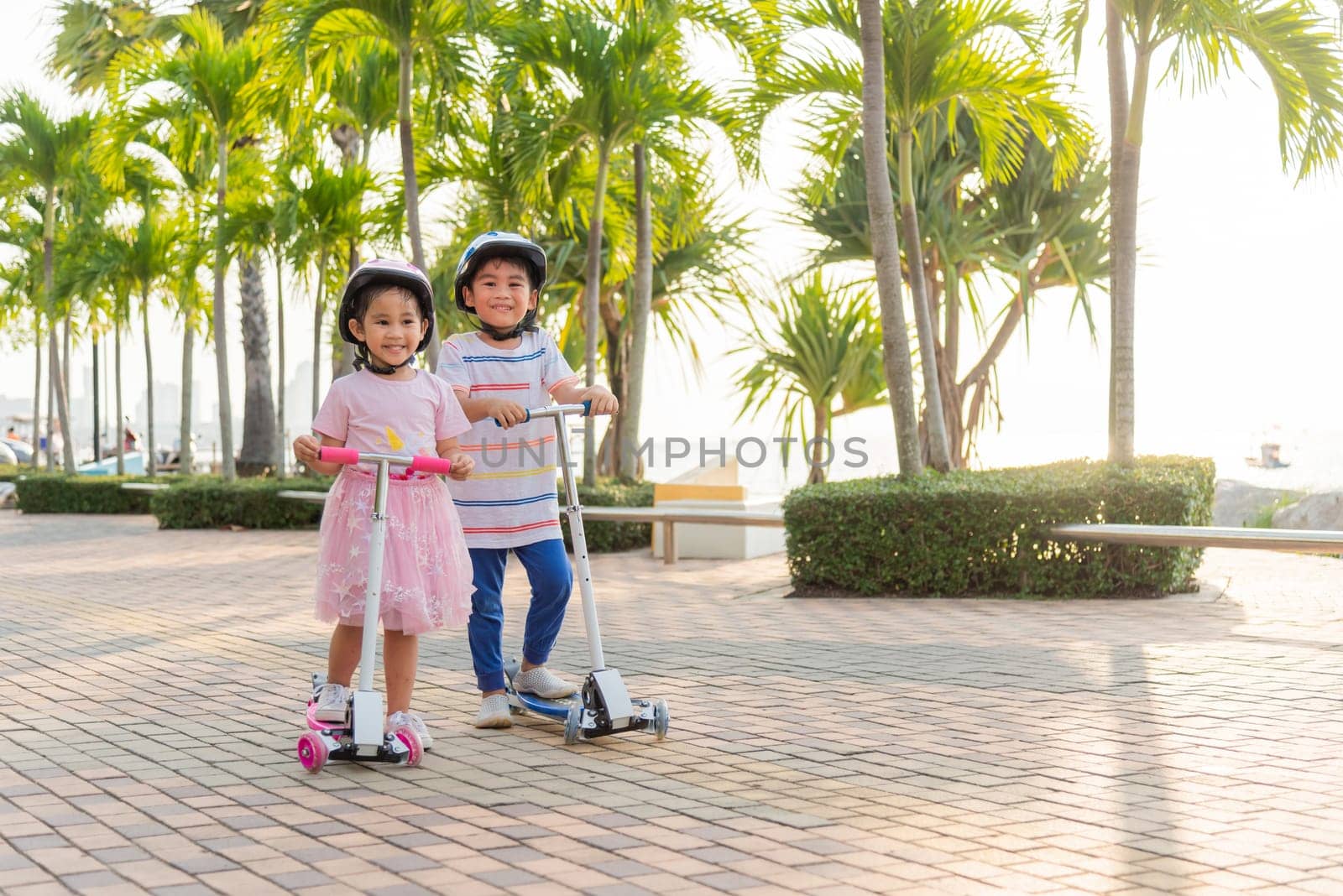 Child riding scooter. Happy Asian little kid boy and girl wear safe helmet play kick board on road in park outdoors on summer day, Active children games outside, Kids sport healthy lifestyle concept