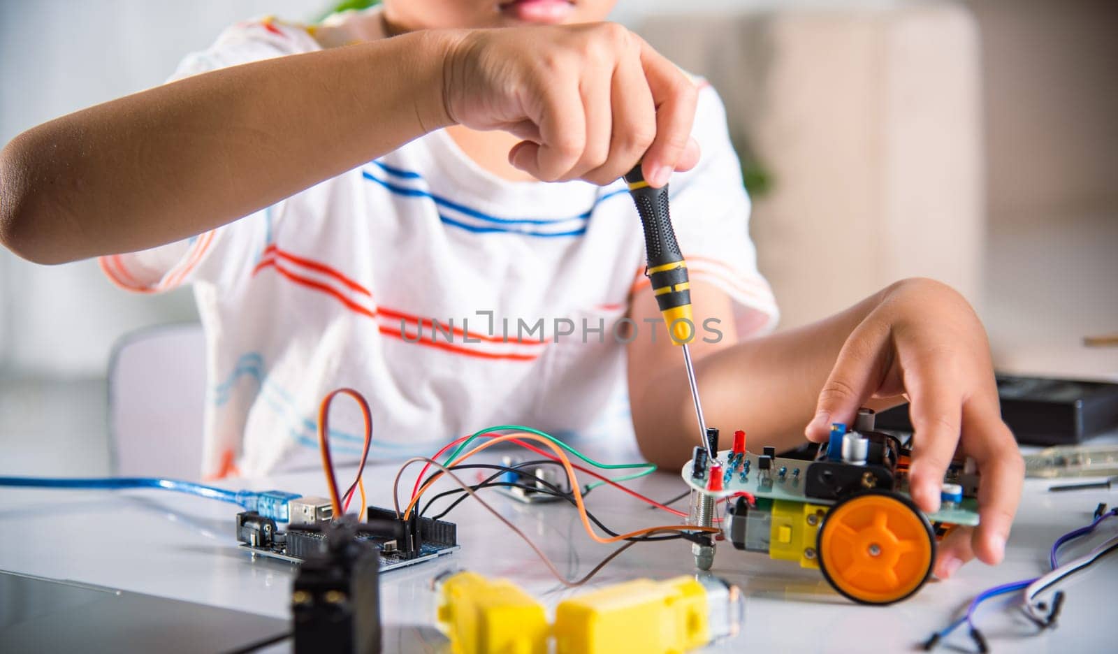 Asian kid boy assembling the Arduino robot car homework project at home, Little child tighten the nut with a screwdriver to assemble car toy, creating electronic AI technology workshop school lesson