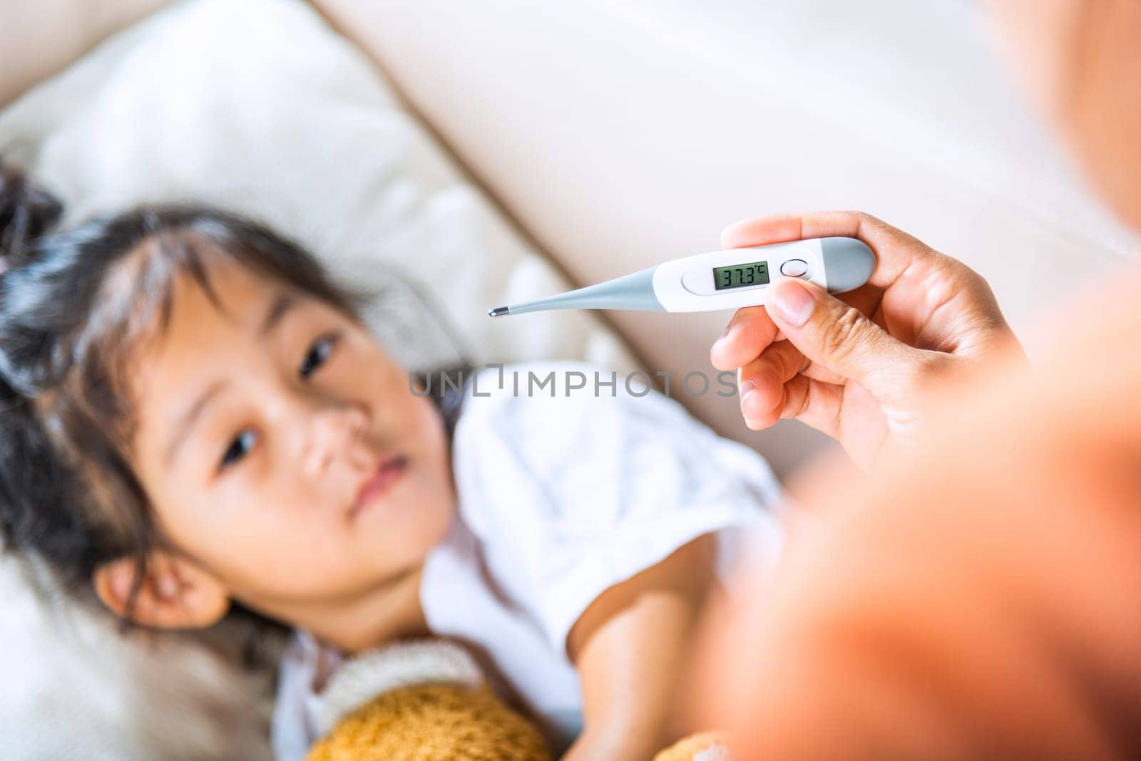 Sick kid. Mother checking temperature of her sick daughter with thermometer in mouth, child laying in bed taking measuring her temperature for fever and illness