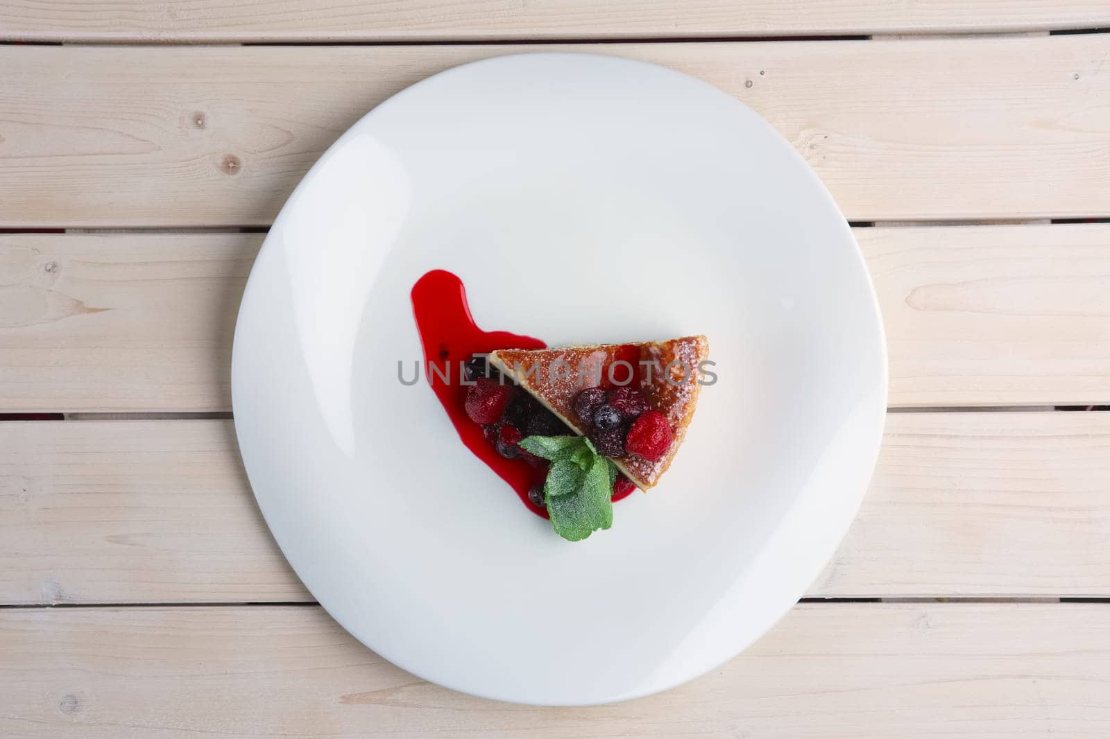 Top view of cheesecake with berries, sauce and green mint leaf on plate on wooden table.