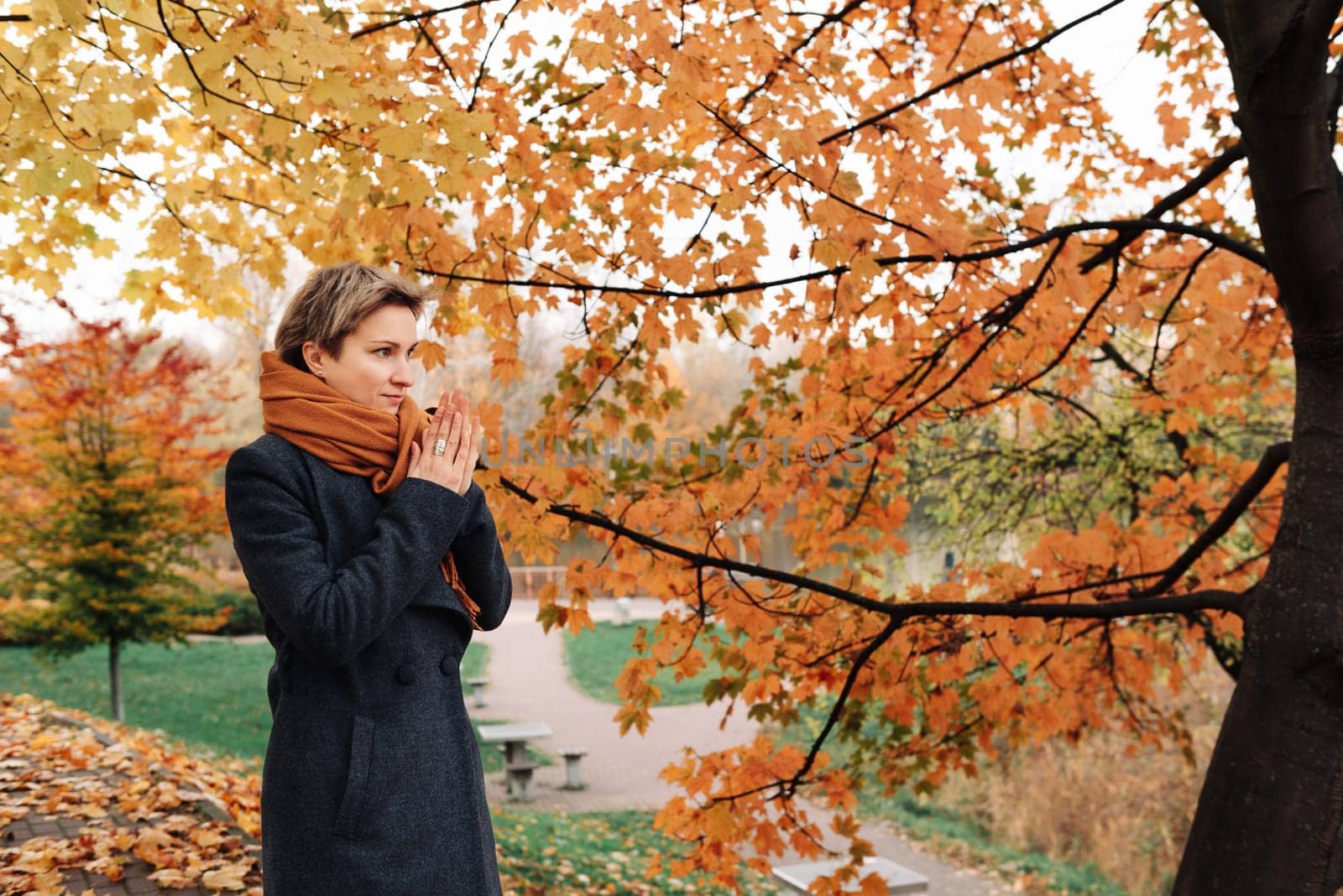 Short-haired blonde in coat and scarf warms her hands in the park.