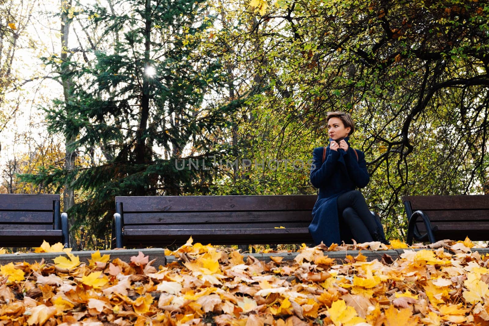 A woman wraps herself in a coat sitting on a bench in an autumn park.