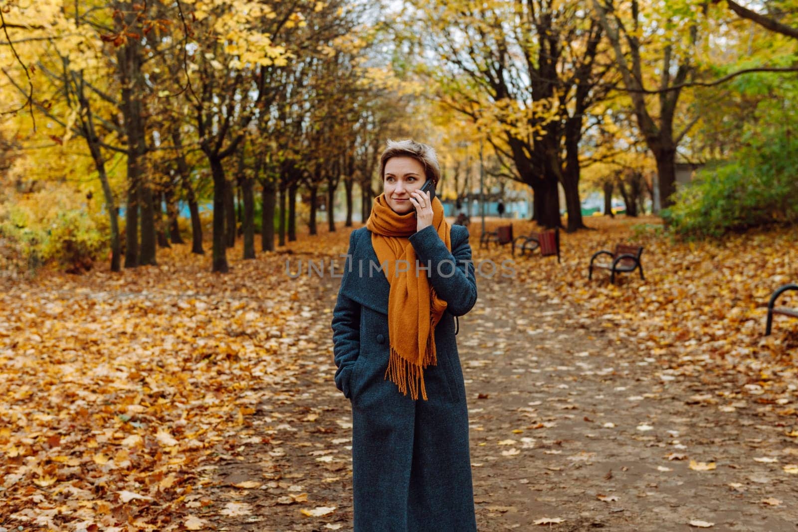A blonde in a coat and scarf talks on the phone as she walks through an autumn park