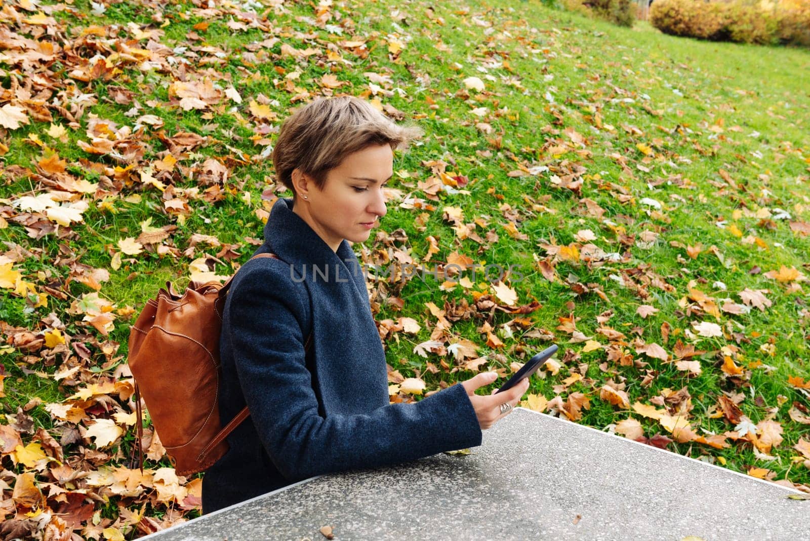 A woman looks at the screen of her phone sitting at a table in an autumn park