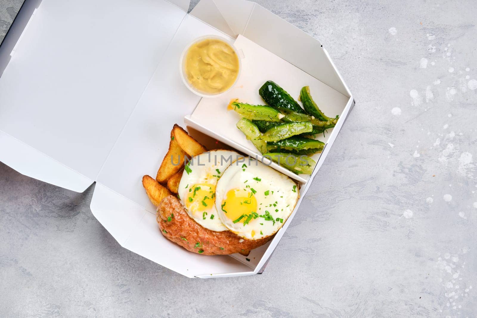 Fried eggs with sausage, potato wedges and pickled cucumber in take away cardboard package