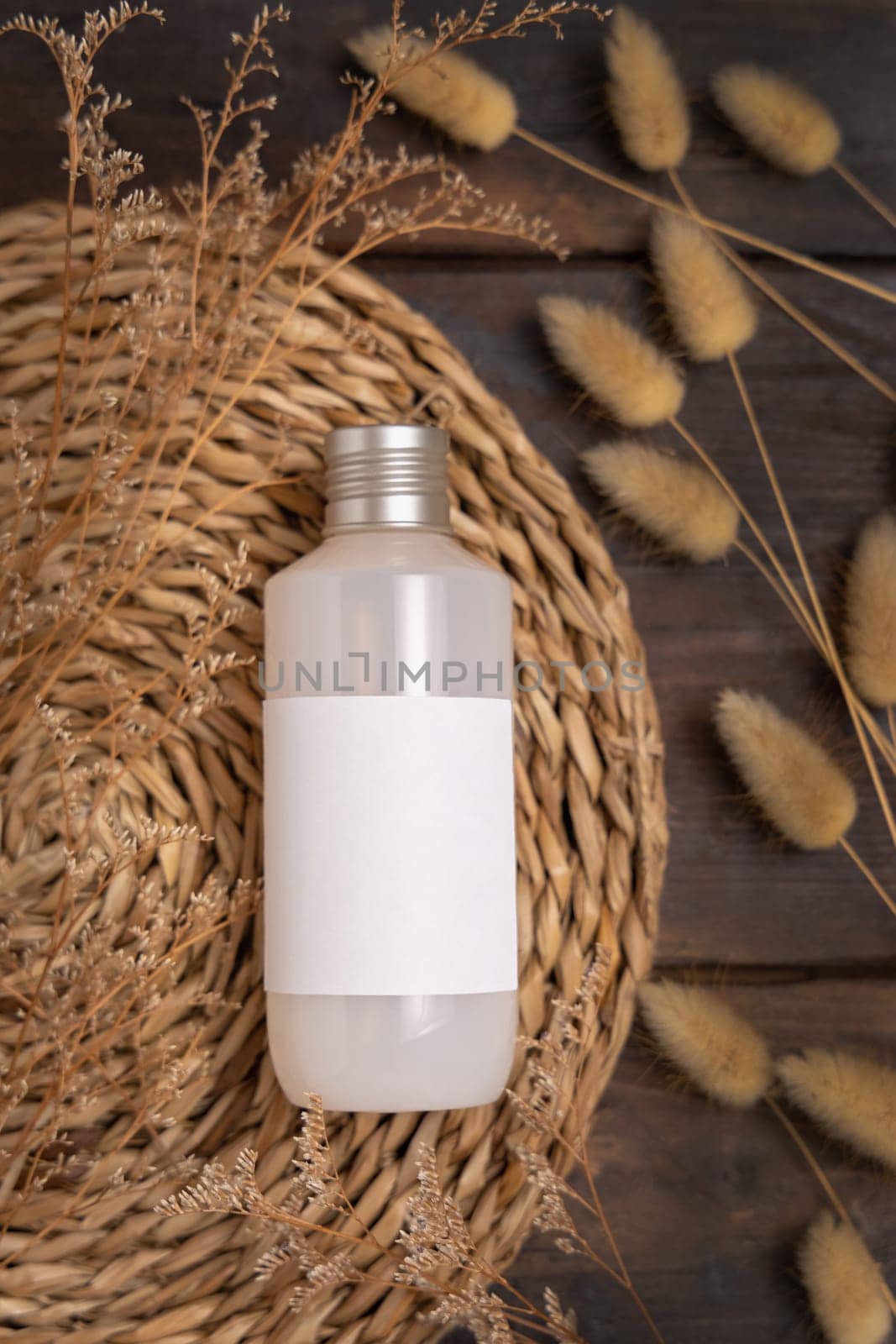 Cosmetic bottle on boho wattled placemat near Hare's Tail grass top view on wood, label mockup. Natural skincare product, cream or lotion. Bohemian eco friendly beauty flat lay.